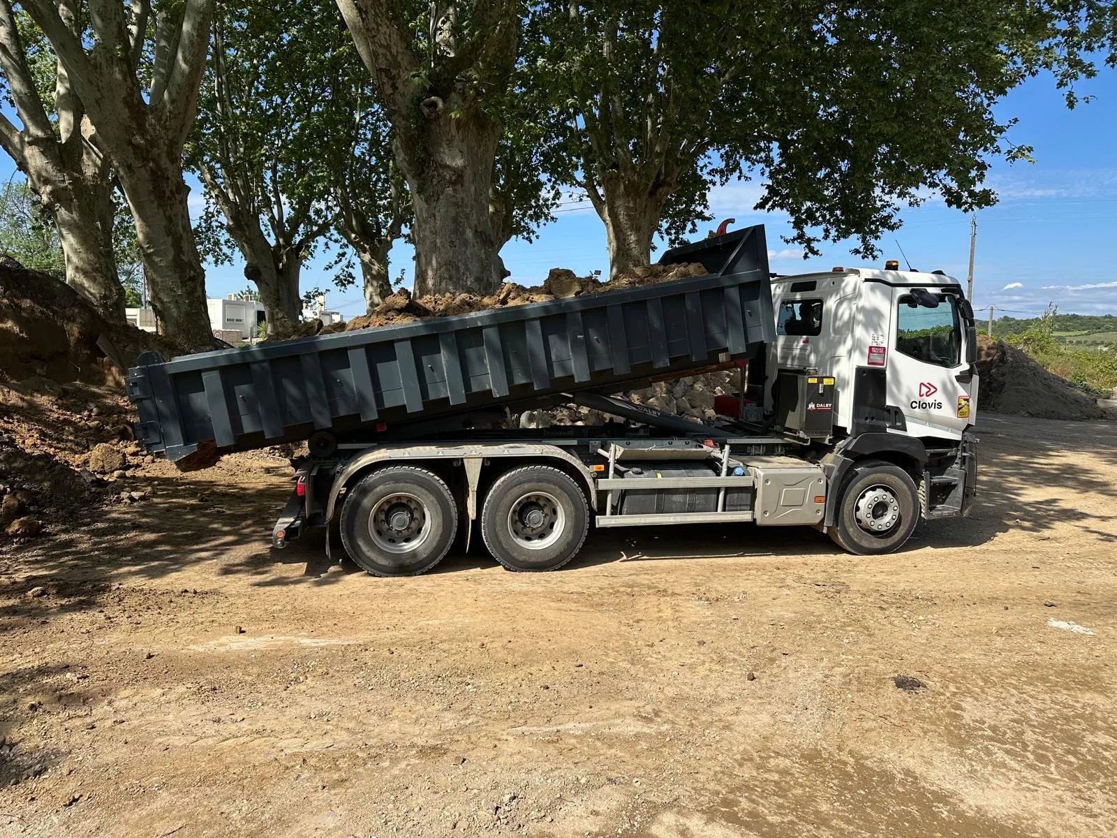 Camion-benne chargé de terre stationné sur un terrain en travaux, sous une rangée d’arbres.
