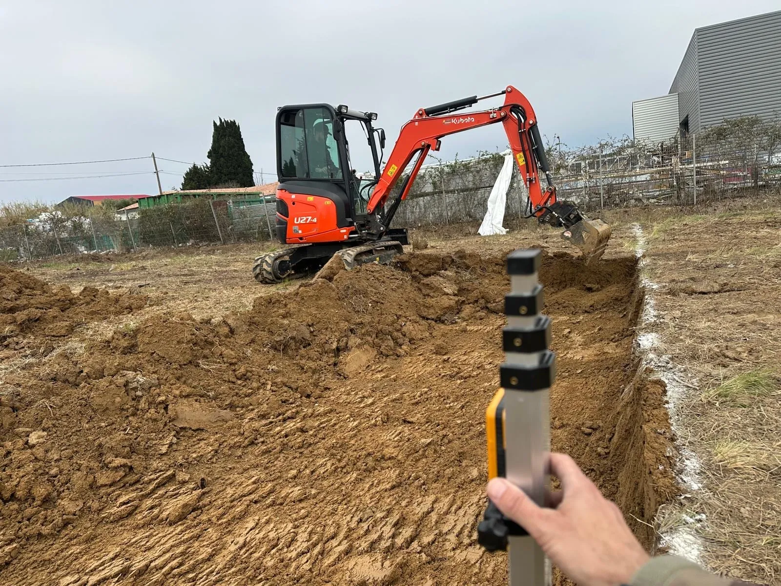 Mini-pelle Kubota creusant une tranchée sur un terrain avec une mire de mesure au premier plan