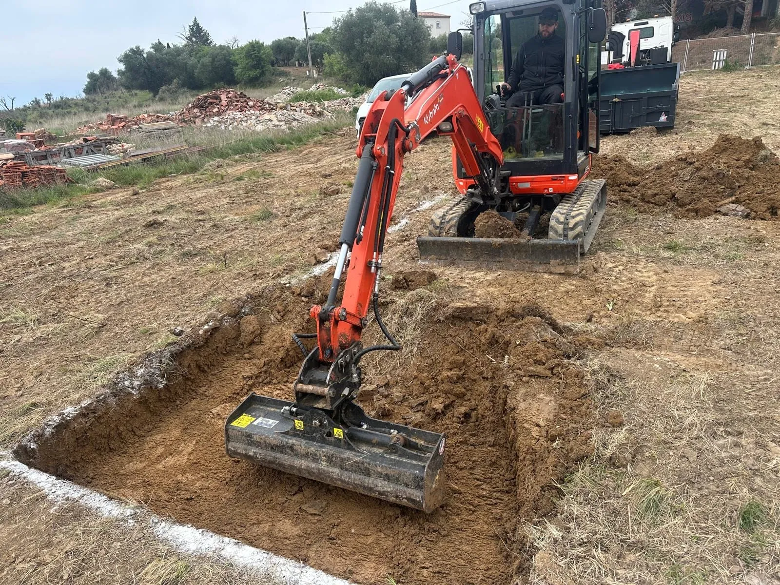 Mini-pelle Kubota creusant une tranchée sur un terrain, avec des gravats et une benne en arrière-plan