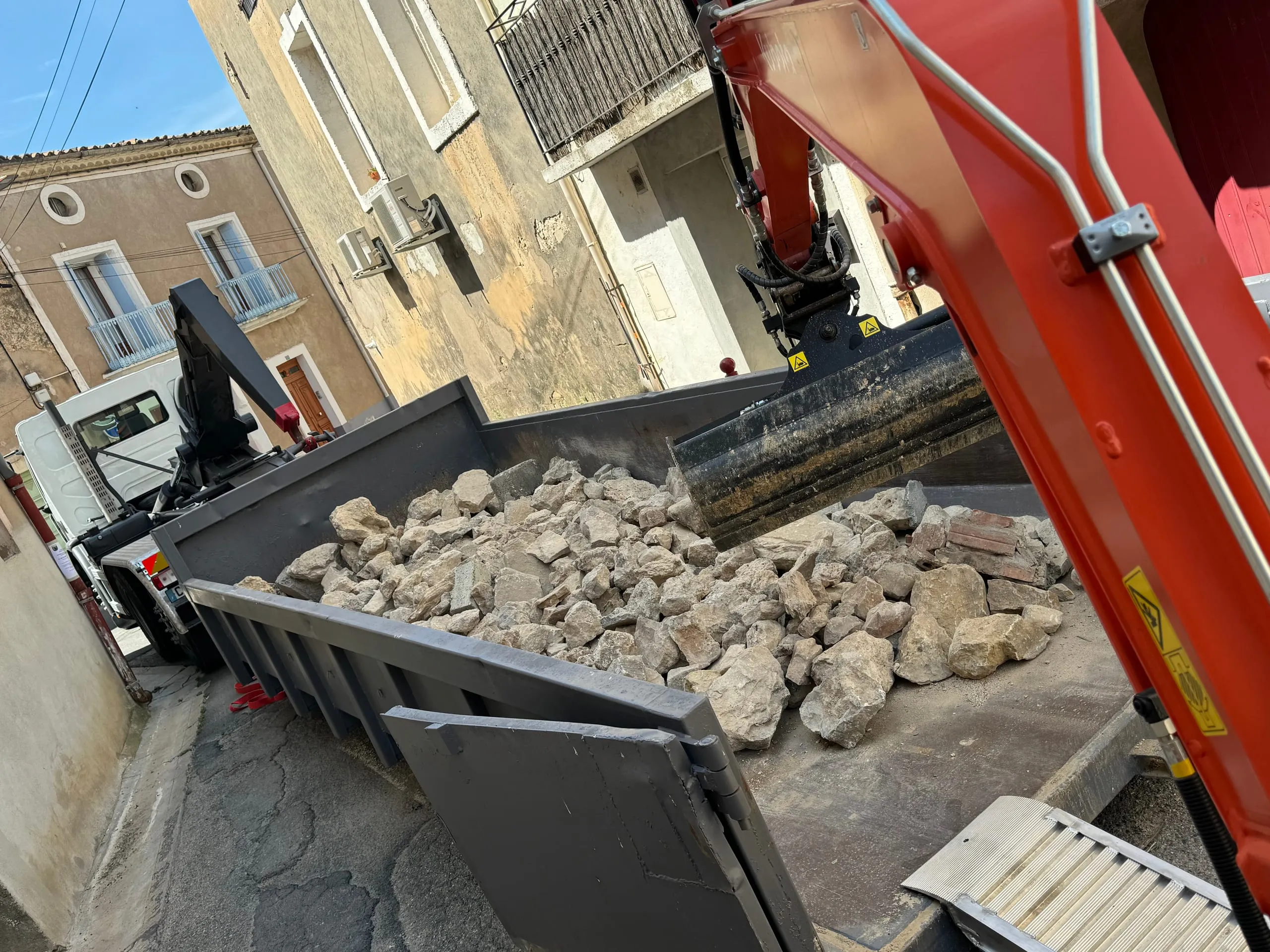 Pelle mécanique chargeant des gravats dans une benne au milieu d’une rue bordée de maisons.