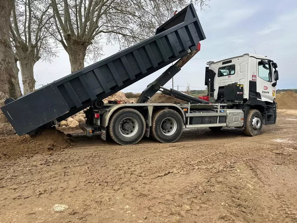 Camion porte-benne blanc déposant une grande benne noire sur un terrain de chantier
