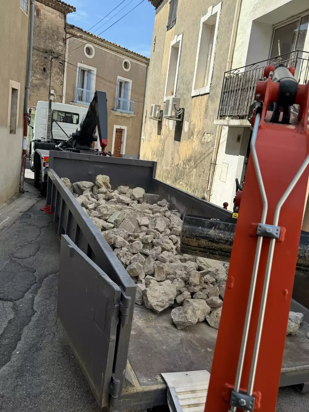 Benne chargée de blocs de pierre avec un bras de mini-pelle dans une rue étroite.