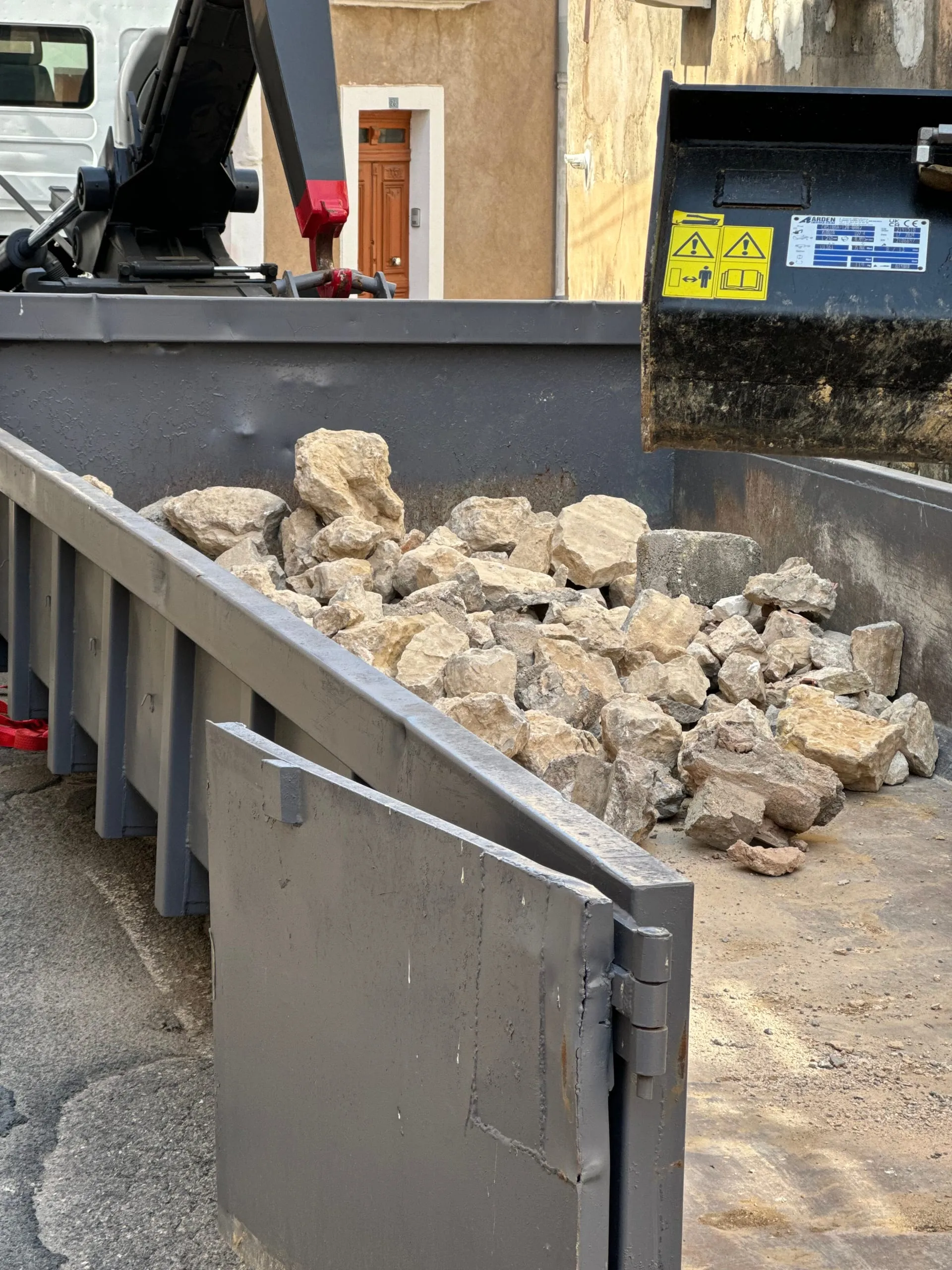 Benne métallique chargée de blocs de pierre devant une maison