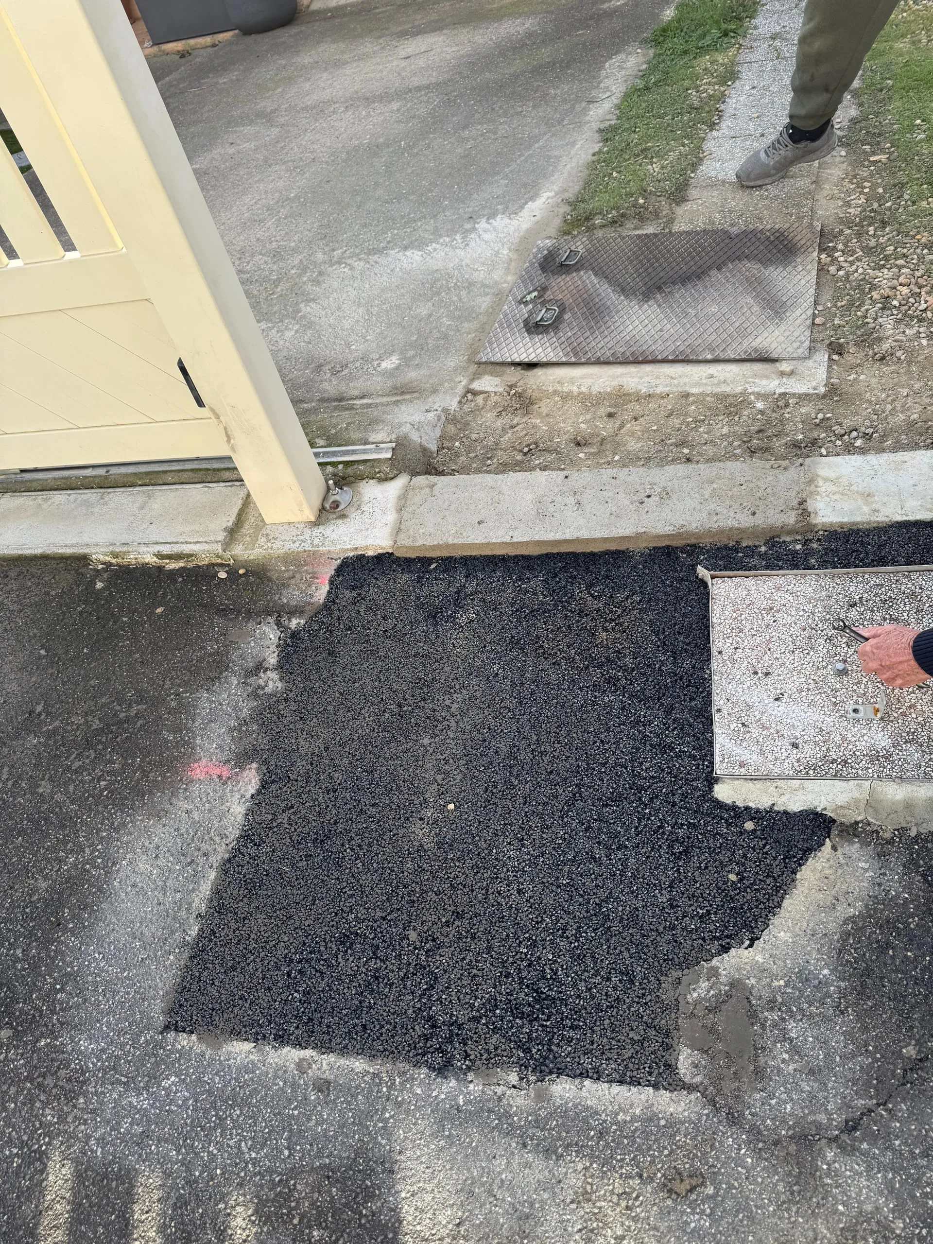 Enrobé noir fraîchement posé devant un portail, entre deux plaques de regard